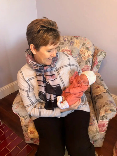 Person in plaid sweater holding baby in coral outfit in floral armchair for pregnancy assistance.